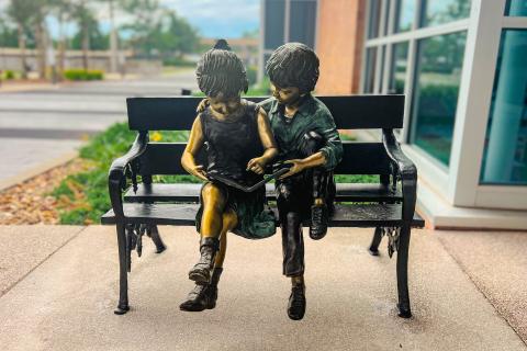 Image of a bronze bench that contains two children sitting on it reading a book 